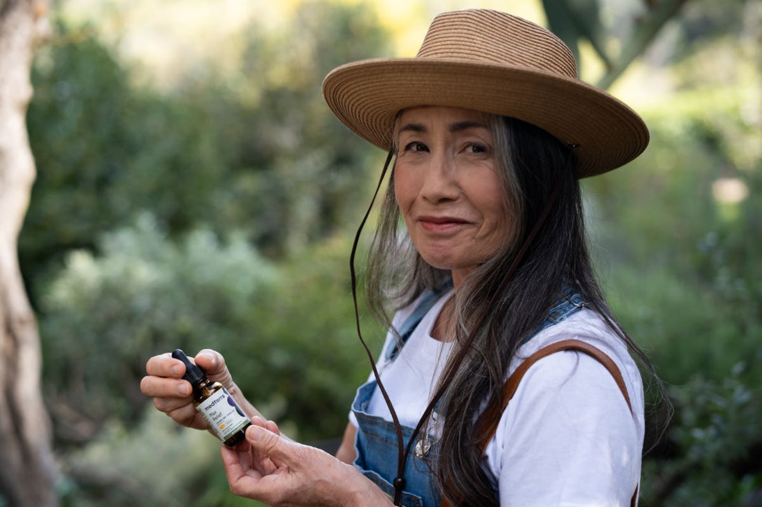 Older woman in a hat holding a Medterra CBD oil dropper bottle outdoors, smiling slightly