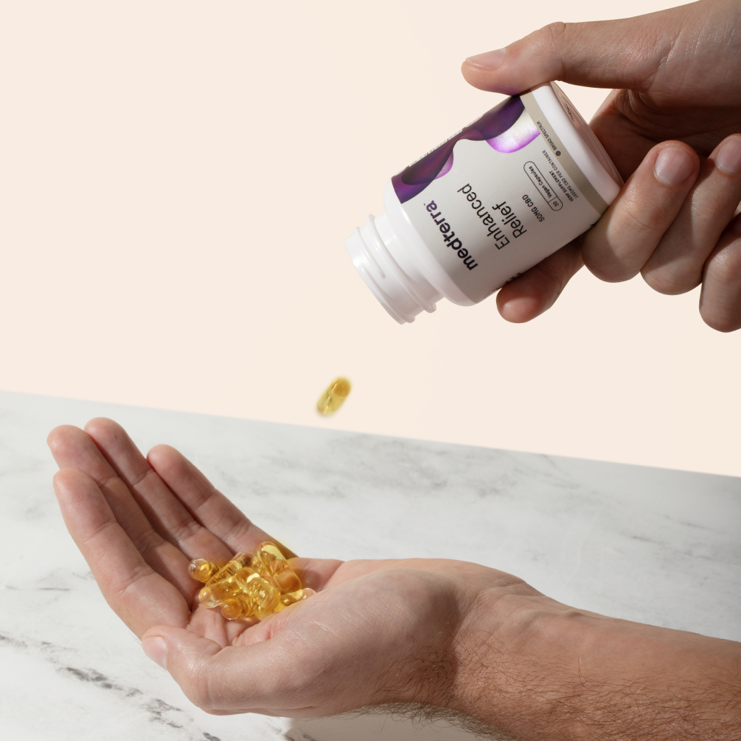 A person's hand is holding an open bottle of Medterra Enhanced Relief Broad Spectrum Capsules, pouring yellow capsules into their other hand against a light background. The bottle has a white label with purple accents.