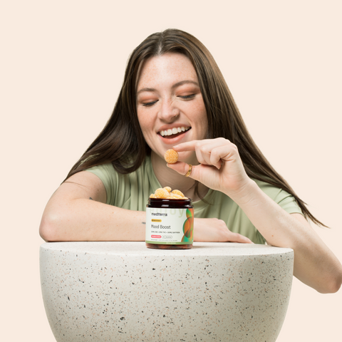 A woman with long hair smiles while holding a Medterra CBD gummy above an open jar labeled "Mood Boost Gummies," filled with more gummies. She leans on a speckled, beige-colored surface against a light background.