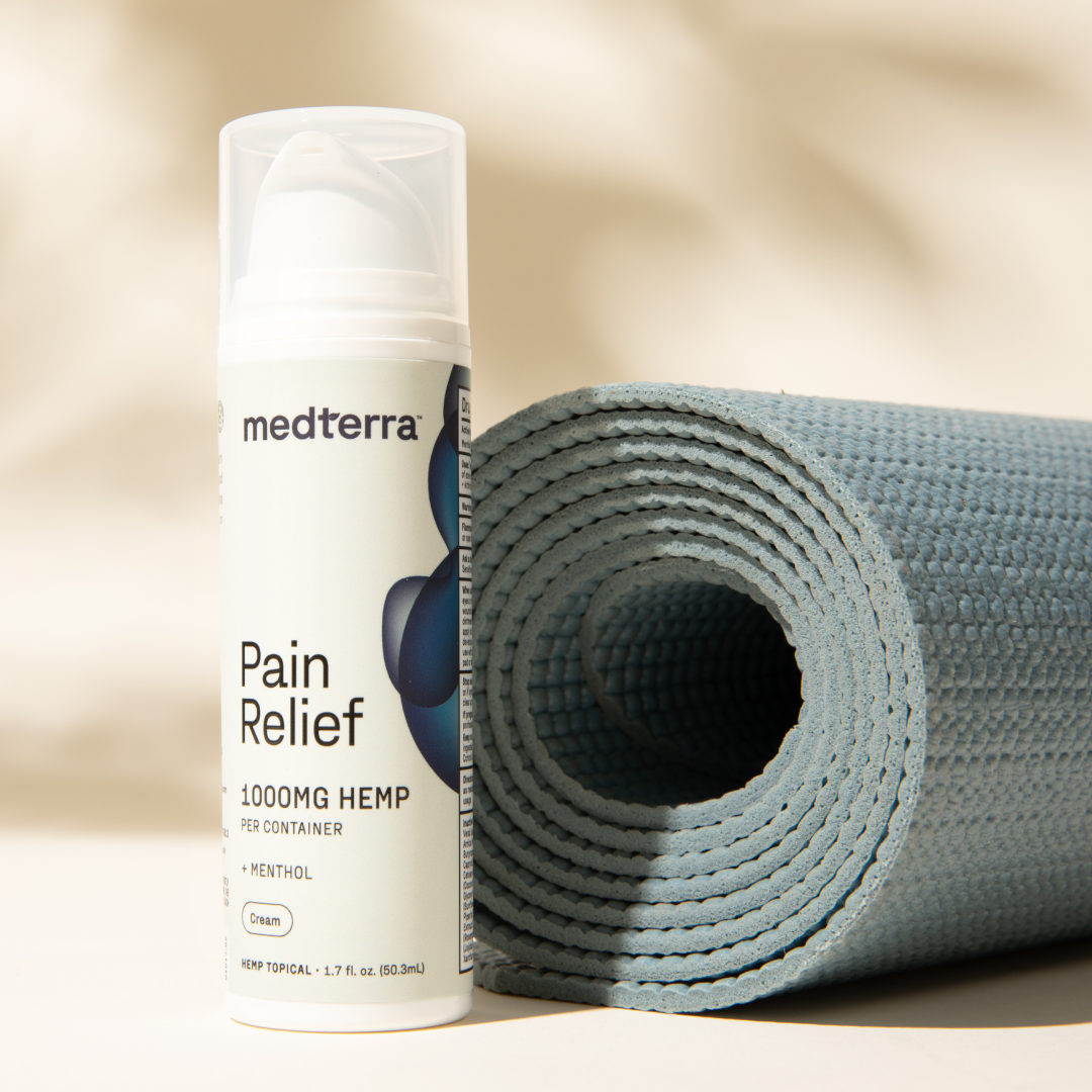 A bottle of Medterra Pain Relief Cream stands upright next to a rolled-up light blue yoga mat, perfect for arthritis and joint pain relief. The label indicates it contains menthol and measures 1.7 fl oz (50 ml). The soft beige background adds warmth to the scene, highlighting this essential CBD personal care product from Medterra.
