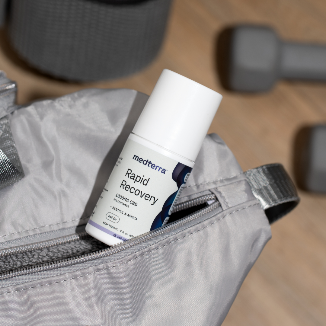 A bottle of Medterra’s Rapid Recovery CBD Roll On, acclaimed for its joint and muscle support along with a rapid cooling effect, is partially zipped inside a gray gym bag. In the background are gray dumbbells and a yoga mat, indicating a fitness or workout environment.