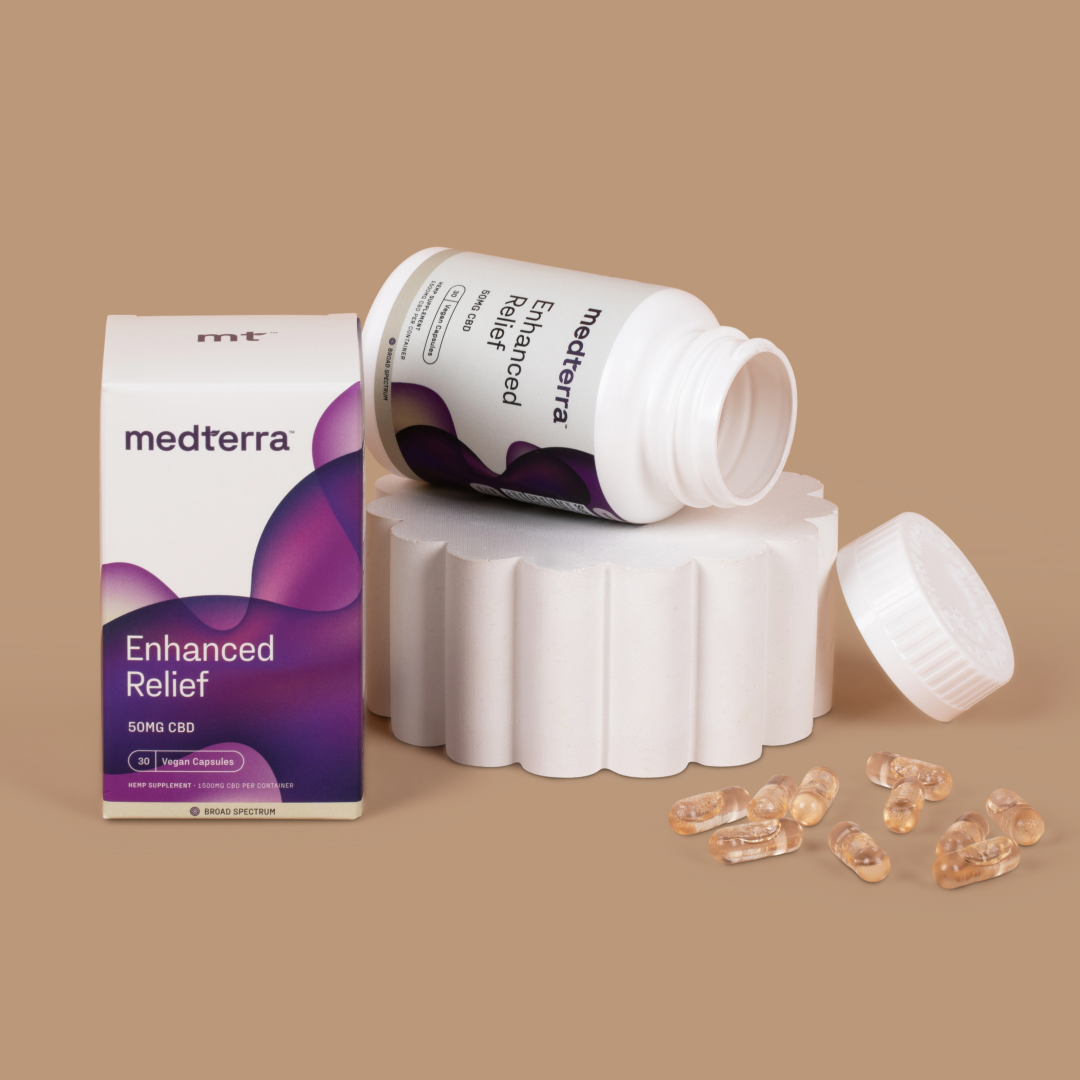 A bottle of Medterra Enhanced Relief Broad Spectrum Capsules (50mg) is shown open, with several capsules spilled out next to it. The bottle and capsules are displayed on a white pedestal, with a branded box and capsule label visible, set against a beige backdrop.