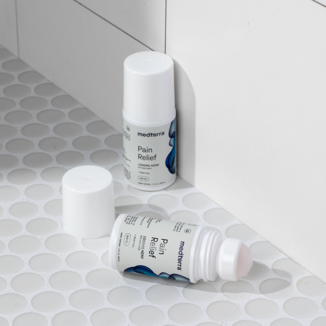 Two Medterra Pain Relief Roll-On bottles, each containing 1000mg of CBD, are displayed against a white tiled surface. One stands upright with its cap on, while the other lies on its side with the cap off, exposing the roll-on applicator crafted for joint pain relief and enduring comfort.