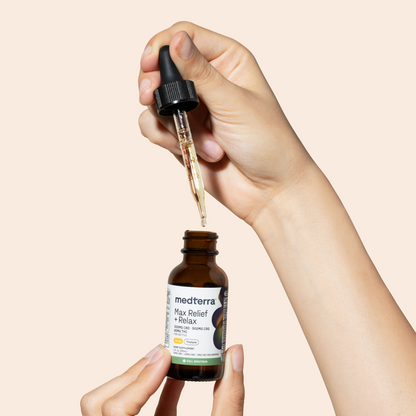 A person is holding a dropper and a bottle labeled "Medterra Max Relief + Relax Full Spectrum CBD Oil" in their hands. One hand holds the brown bottle of the Max Relief + Relax Full Spectrum CBD Oil while the other hand holds a dropper filled with the liquid product, ready to dispense for optimal relaxation support.