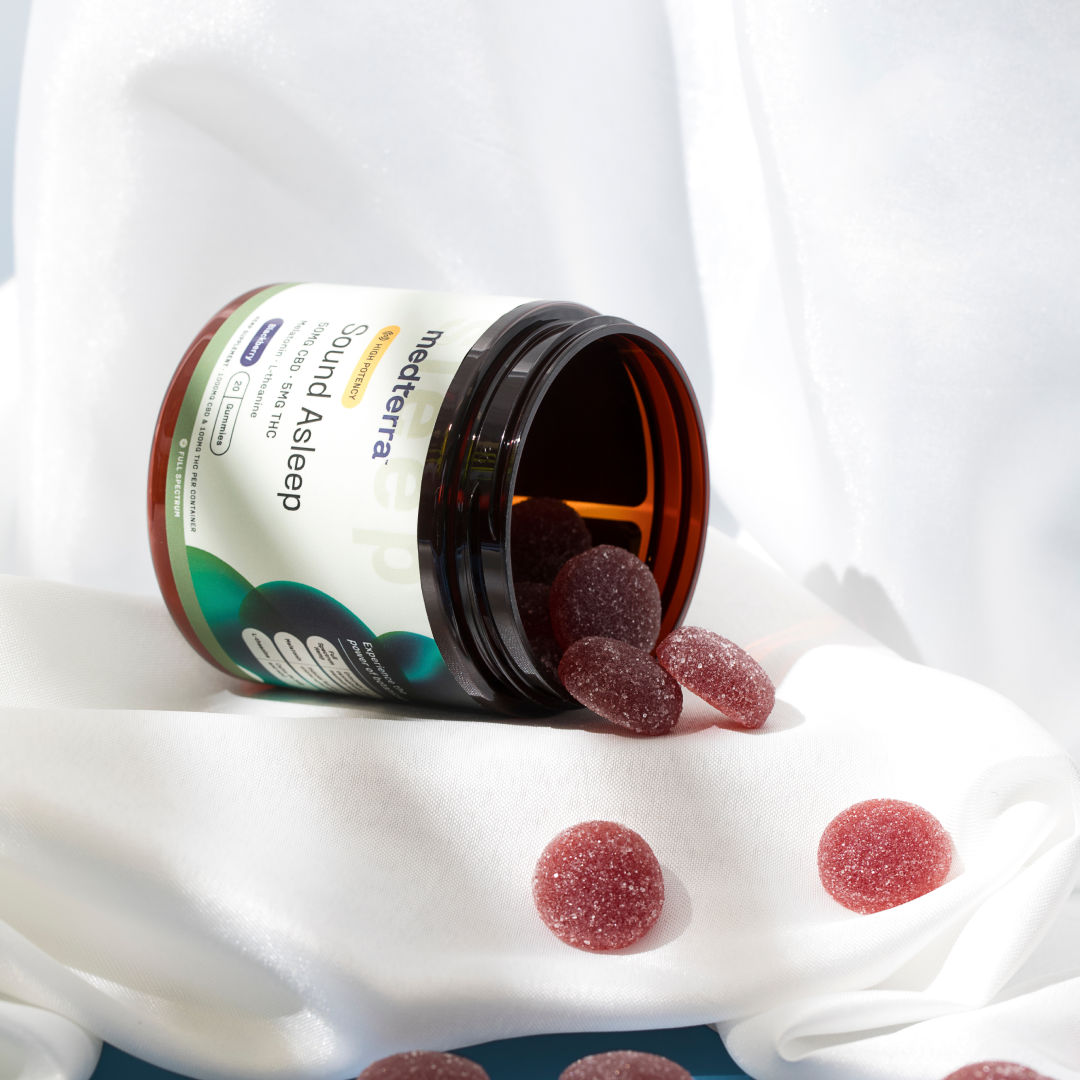 An open jar of Medterra Sound Asleep Gummies, a reliable sleep aid, with several dark red gummies spilling out onto a white cloth. The jar and gummies are softly illuminated, with a backdrop of white, flowing fabric.