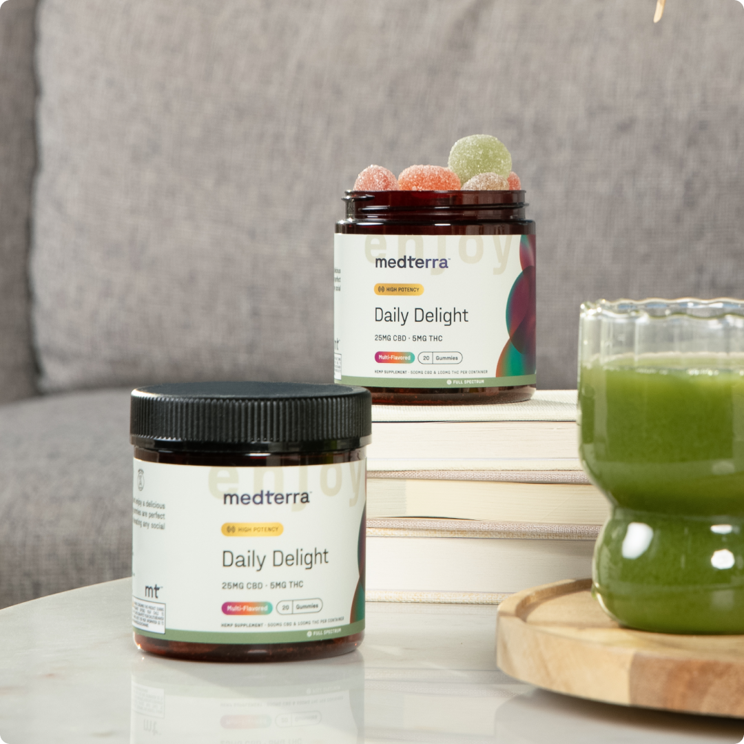 Two jars from the Medterra Daily Delight Bundle rest atop stacked books next to a green drink on a wooden tray, with a blurred gray couch in the background.
