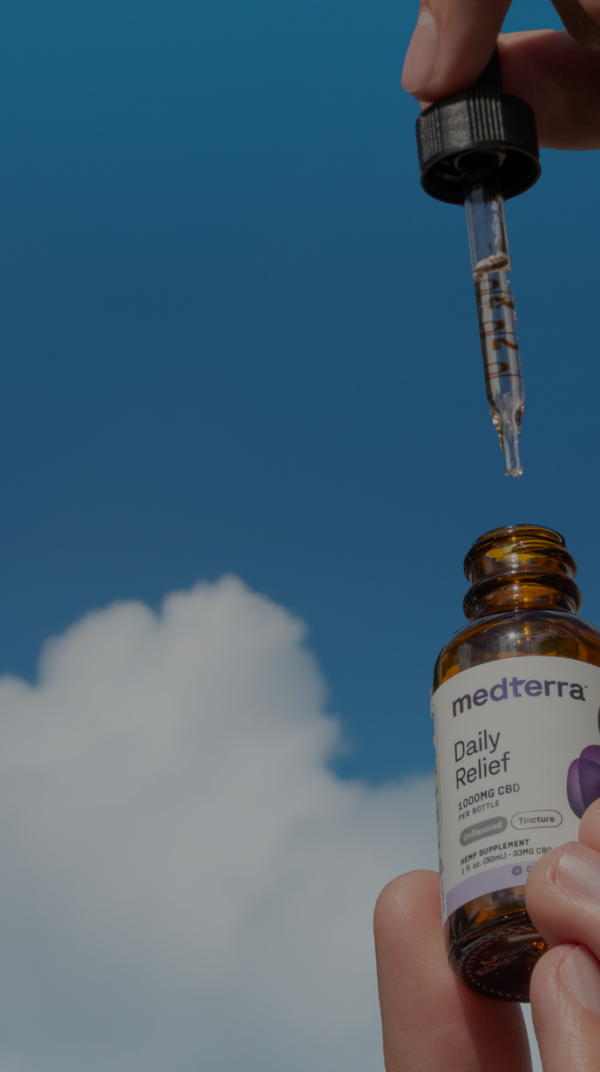 Hand holding medterra bottle labeled 'Daily Relief 1000MG CBD PER BOTTLE Unflavored Tincture' with dropper against blue sky