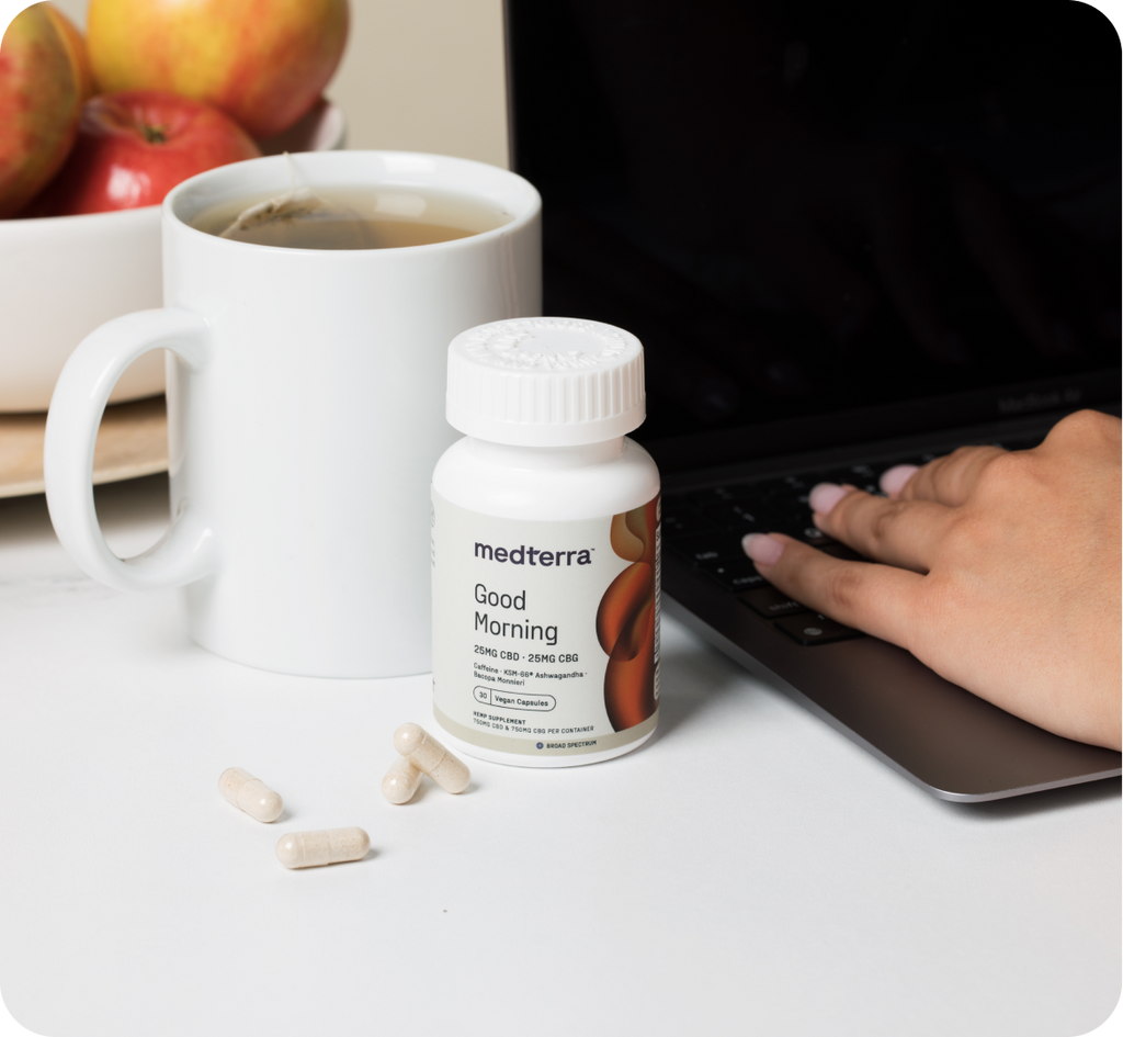 A person types on a laptop next to a white mug, a bottle labeled "medterra Good Morning," and several vegan capsules spread on the table. In the background, a bowl of apples is partially visible, setting the scene for an energized morning routine.