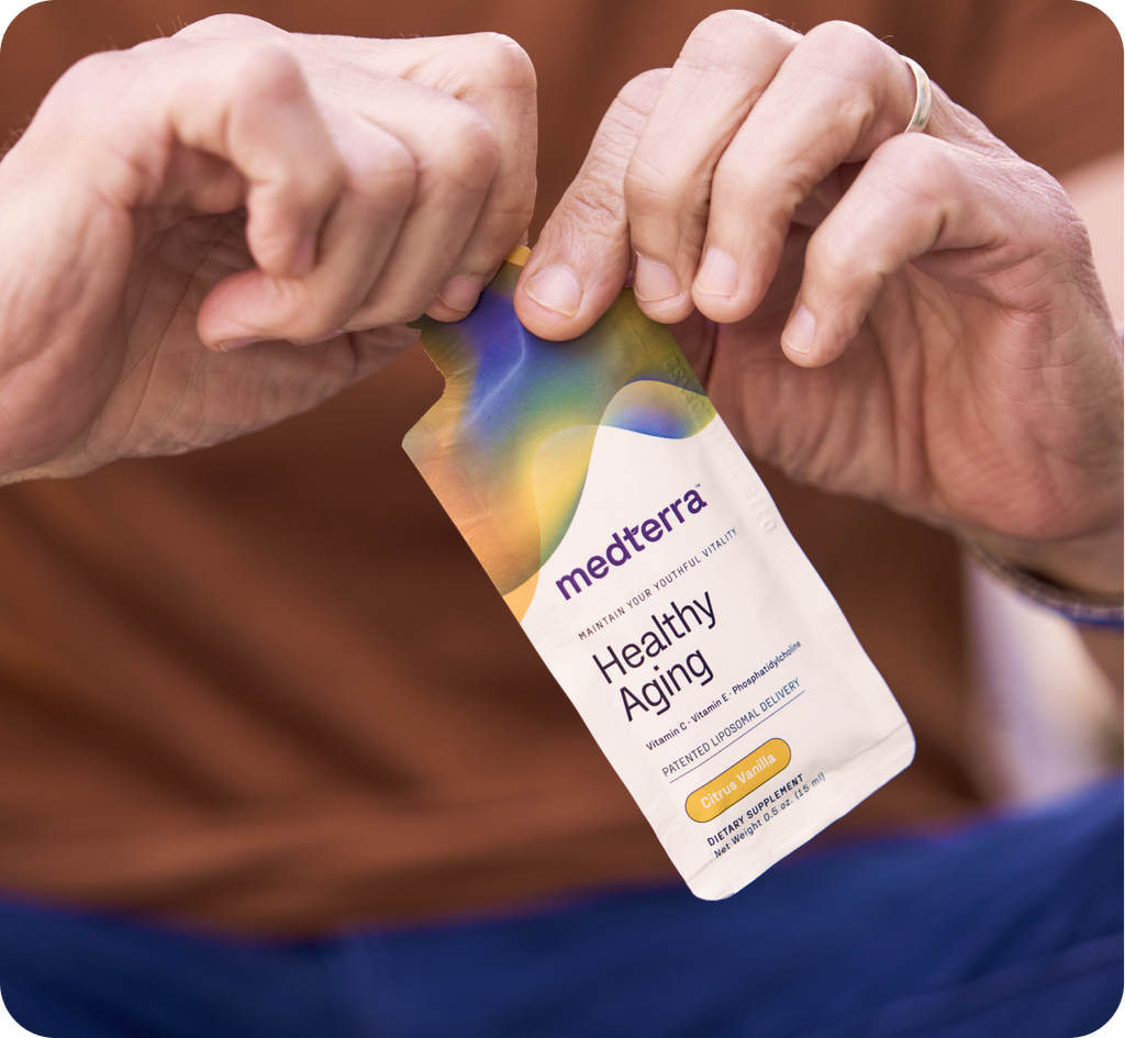 A person is holding and tearing open a packet labeled "medterra Healthy Aging" in their hands. The colorful gradient design is eye-catching and indicates it contains Citrus Sunset flavored supplements with liposomal delivery, enriched with Vitamin C to support your healthy aging journey.