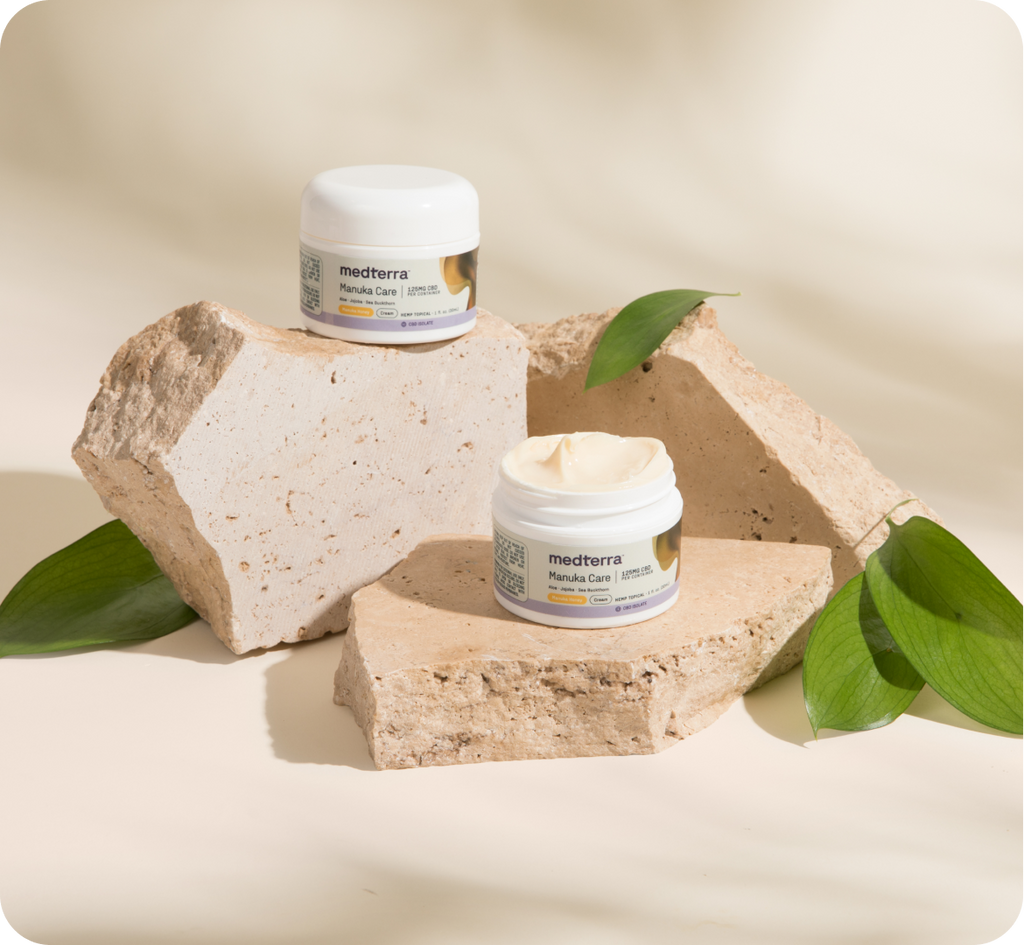 Two jars of Medterra Manuka Cream are placed on beige stone slabs, surrounded by green leaves. One jar has its lid off, revealing the rich CBD skin cream inside. The neutral beige background enhances the natural aesthetic.