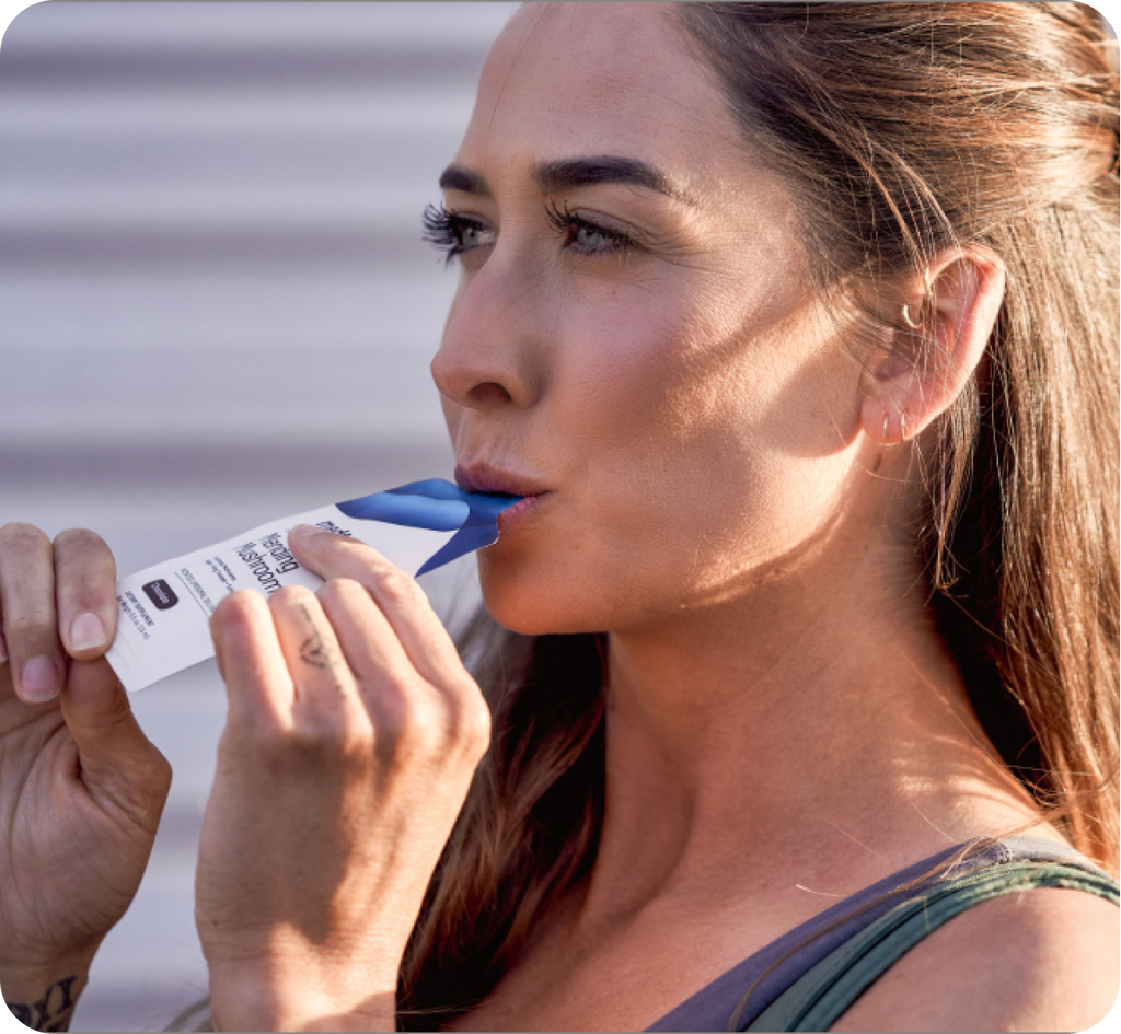 A woman with long brown hair drinks from a hydration packet outdoors. She is wearing a grey tank top and has a slight smile on her face. The packet, held in her hand, features Mending Mushrooms with antioxidant benefits and a blue spout that she is sipping from. Sunlight illuminates her face from the side.