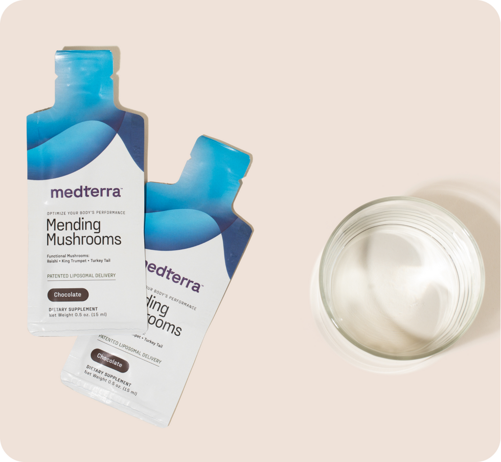 Two Medterra sachets labeled "Mending Mushrooms" with a chocolate flavor sit beside a clear glass of white liquid on a neutral backdrop. The blue and white packaging highlights the functional mushroom benefits and antioxidant benefits, ideal for those seeking a vegan formula solution.