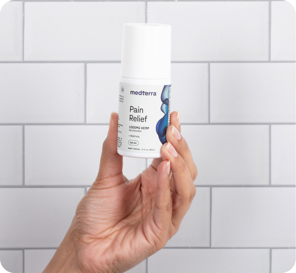 A hand is holding a cylindrical bottle of Medterra Pain Relief Cream against a white tiled background. The bottle has a white lid and label, with blue accents, indicating it contains 1000mg of CBD with arnica and menthol – an ideal solution for arthritis pain relief.