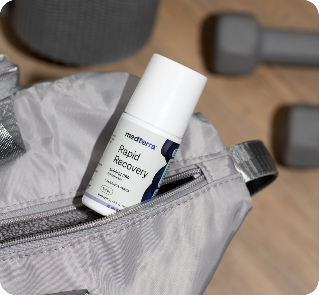 A Medterra Rapid Recovery roll-on bottle, known for its joint and muscle support, is partially visible inside a gray, open gym bag. In the background, two dumbbells and a cylindrical piece of fitness equipment rest on the wooden floor.