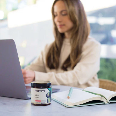 A jar of Medterra Calm CBD product on a desk with a woman working on a laptop in the background.