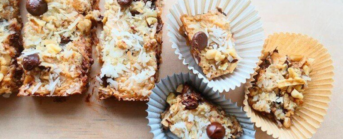 Close-up of creamy coconut cookie bars with chocolate chips and chopped nuts, some placed in white and brown paper liners on a light surface.