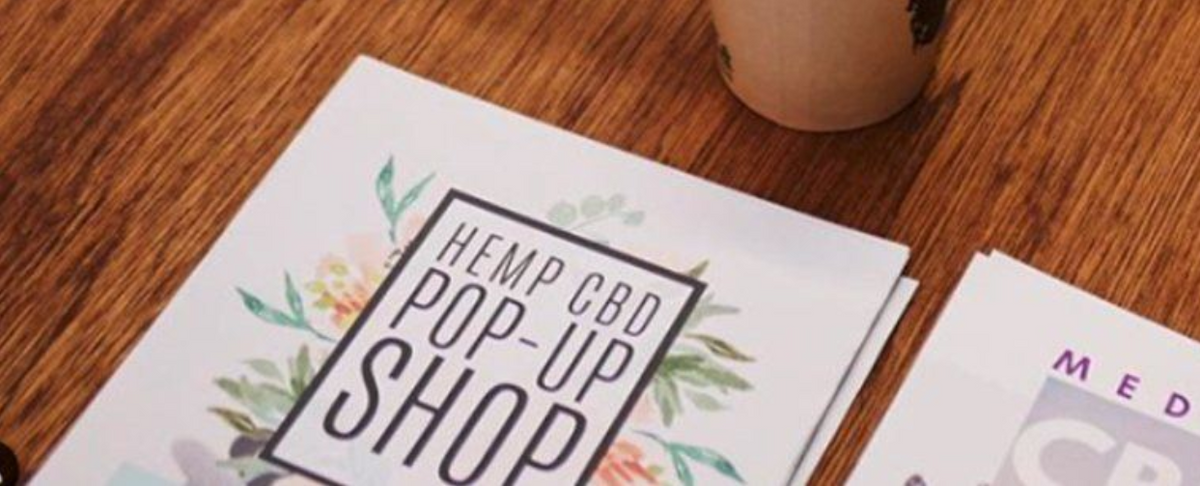 Flyer on wooden table with text 'HEMP CBD POP-UP SHOP' surrounded by floral decoration.
