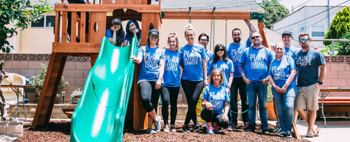 Group of people wearing blue shirts with text 'PLAY DEFEATS CANCER' and one person wearing a shirt with 'BUILD HOPE', standing around a green playground slide outdoors.