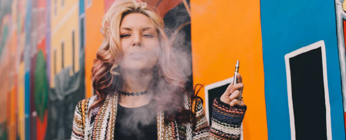 Woman exhaling vapor from a CBD vape pen in front of colorful urban mural walls