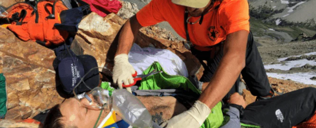 A rescuer wearing gloves and an orange shirt providing emergency medical aid to a person lying on rocky terrain with an oxygen mask and neck brace, suggesting a mountain rescue scenario.