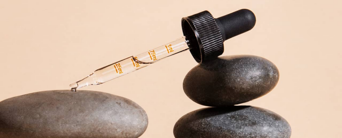 CBD oil dropper releasing a droplet over stacked smooth stones on a beige background, glass pipette showing 0.25–1 ml markings