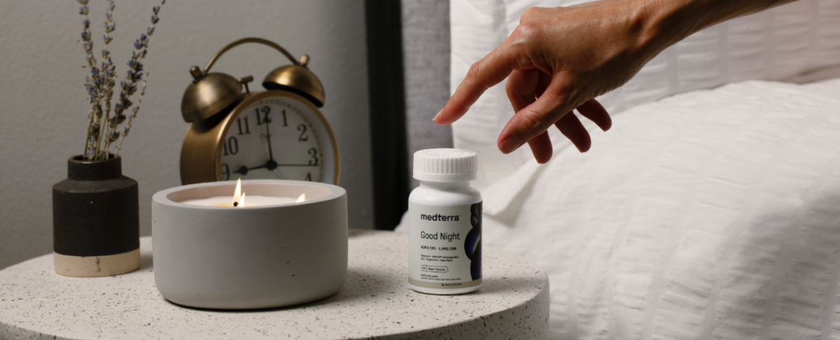 A hand reaching for a bottle of Medterra Good Night CBD capsules next to a lit candle and clock on a bedside table.