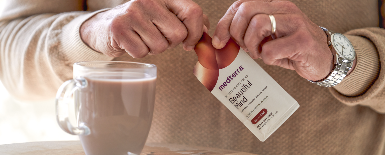 A person wearing a light brown sweater and a silver watch is seen opening a Medterra "Beautiful Mind" supplement packet. In front of them is a glass mug filled with chocolatey liquid placed on a wooden surface.