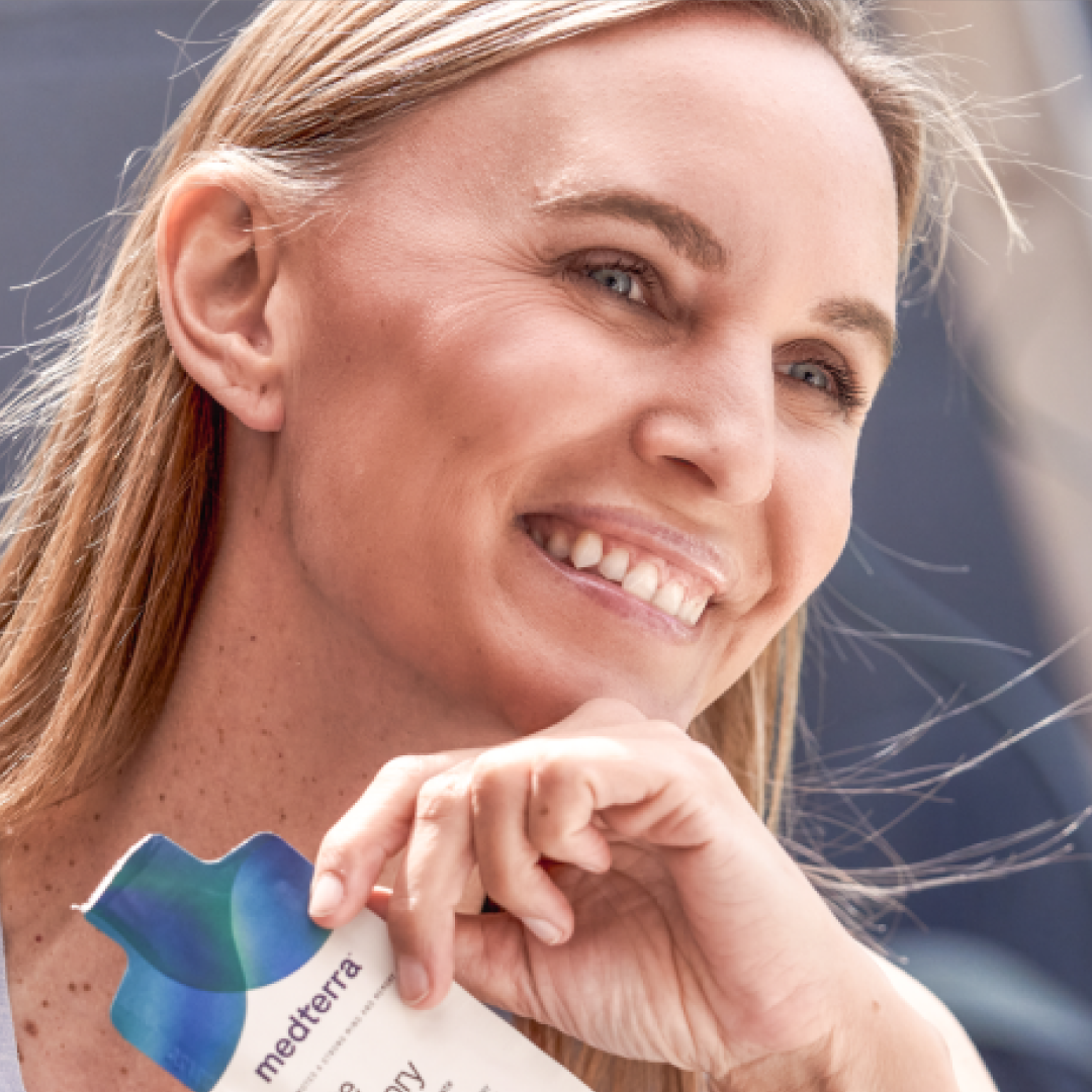 A smiling individual with long, straight hair is holding a Medterra Brain Power product close to their face. They are gazing off into the distance with a content expression, possibly enjoying the advantages of liposomal technology. The background is blurred and indistinct.