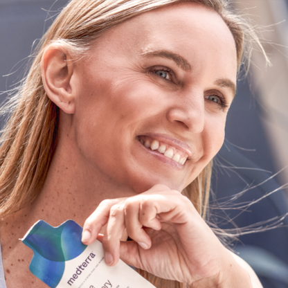 A smiling individual with long, straight hair is holding a Medterra Brain Power product close to their face. They are gazing off into the distance with a content expression, possibly enjoying the advantages of liposomal technology. The background is blurred and indistinct.