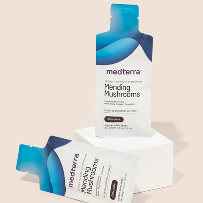 A standing and lying down Medterra Mending Mushrooms supplement packet, designed in shades of blue and white, are displayed against a light background. The standing packet is placed on a white pedestal, showcasing the label and chocolate flavor, emphasizing the mushroom blend's antioxidant benefits ideal for athletic recovery.