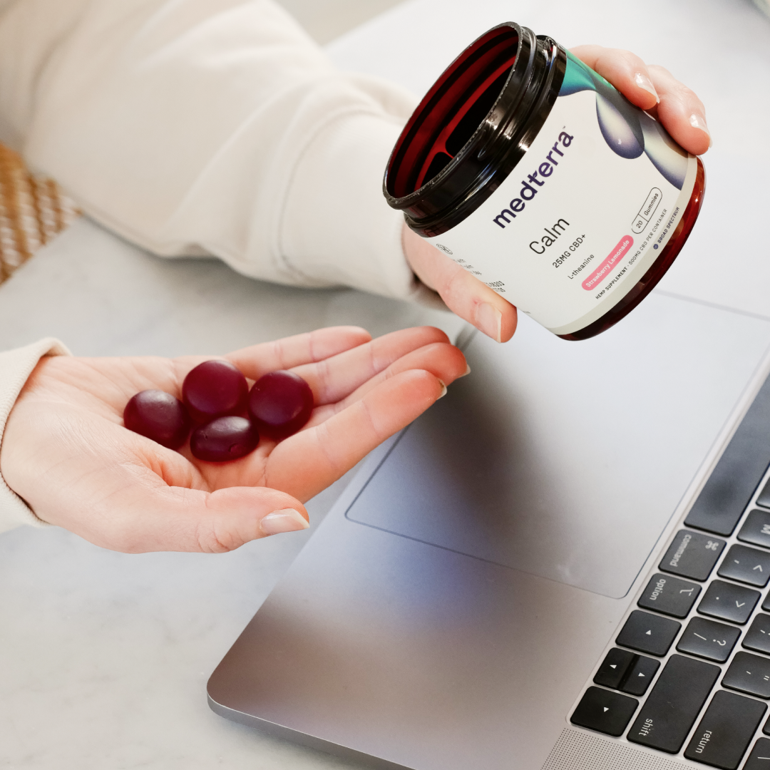 A hand holds out three round, dark red Calm Gummies, while the other hand pours them from an open jar labeled "Medterra." The jar is held over a laptop on a light-colored surface. The person appears to be preparing to consume the broad-spectrum CBD supplements.