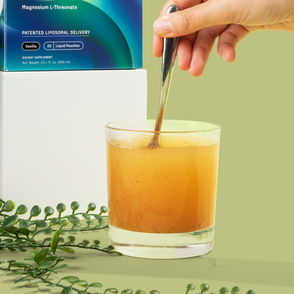 A hand is stirring a yellowish-orange liquid in a glass with a spoon. The backdrop features green leaves and a product box labeled "Brain Power" by Medterra on a white cube, highlighting its vanilla flavor and advanced liposomal technology designed to support brain function.