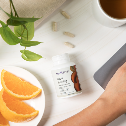 A white table with a bottle of Medterra "Good Morning Capsules" offering cognitive support, a plate of orange slices, a cup of tea, some green leaves, and a hand using a computer mouse. Three CBD-infused capsules are scattered near the bottle.
