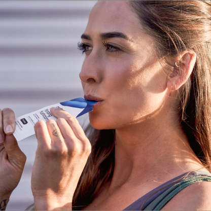 A woman with long hair drinks from a blue and white hydration sachet of Mending Mushrooms by Medterra. She is outdoors, dressed in a tank top, and appears focused. Sunlight casts shadows on her face as she enjoys the antioxidant benefits essential for athletic recovery.