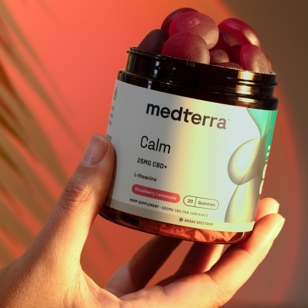 A hand holds a black jar labeled "Medterra Calm Gummies" in white text, containing 20 strawberry lemonade flavored CBD gummies. Each gummy includes 25mg of broad-spectrum CBD and L-theanine for added relaxation. The background features blurred red and yellow hues.