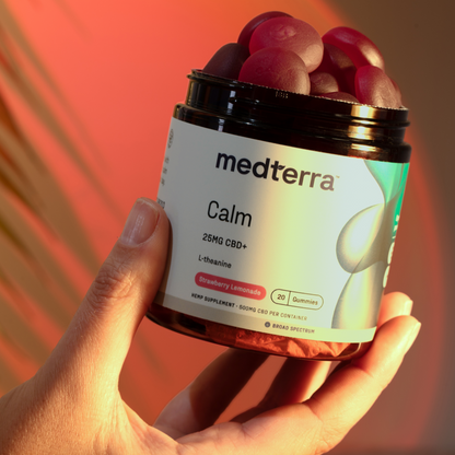 A hand holds a black jar labeled "Medterra Calm Gummies" in white text, containing 20 strawberry lemonade flavored CBD gummies. Each gummy includes 25mg of broad-spectrum CBD and L-theanine for added relaxation. The background features blurred red and yellow hues.