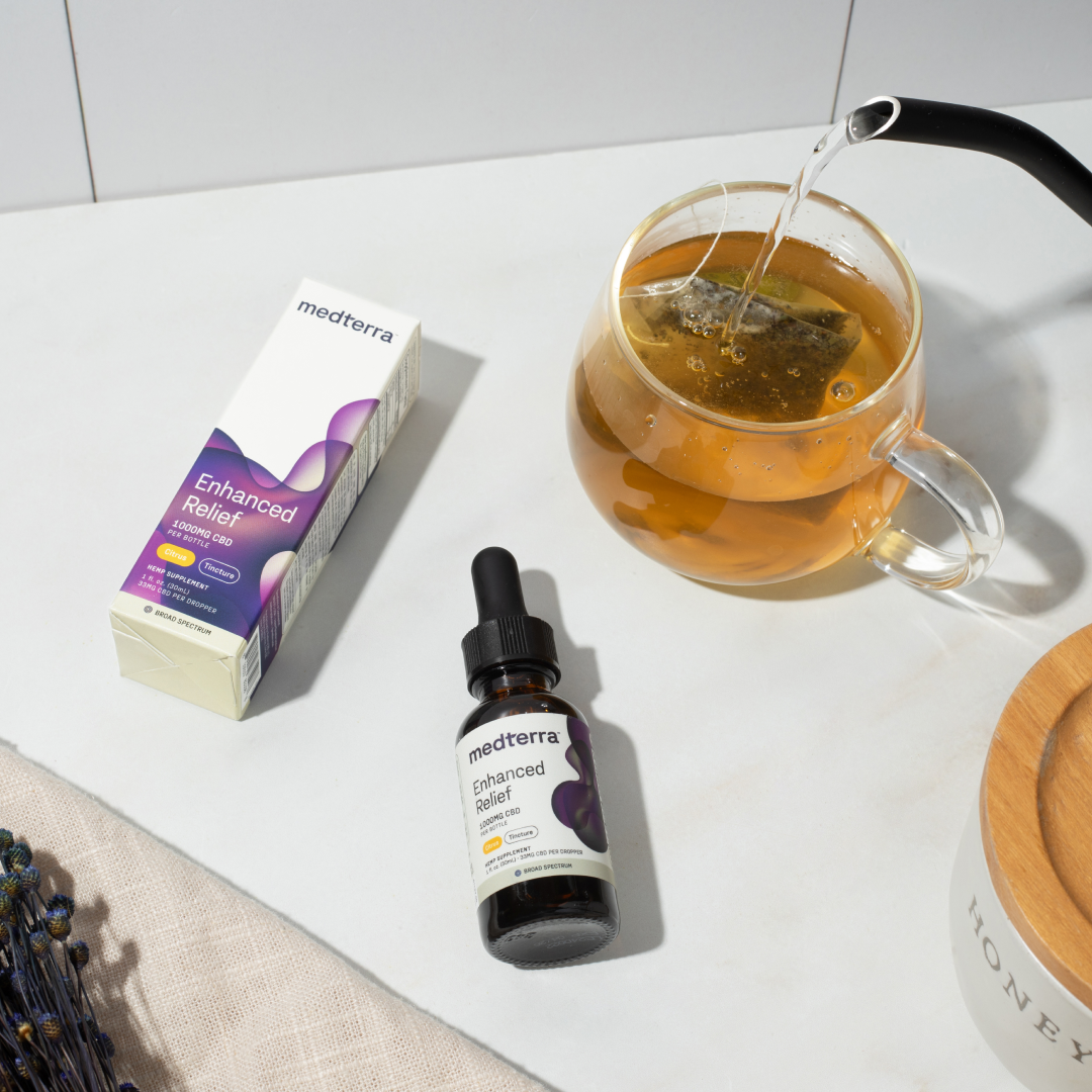 A bottle of Medterra Enhanced Relief Broad Spectrum CBD Oil and its packaging sit on a pristine white marble surface. Nearby, a clear glass teapot holds steeping tea, with hot water being poured from a kettle. The scene is complemented by a cloth and a jar labeled "Honey", highlighting the Broad Spectrum formula which harnesses the entourage effect for optimal relief.