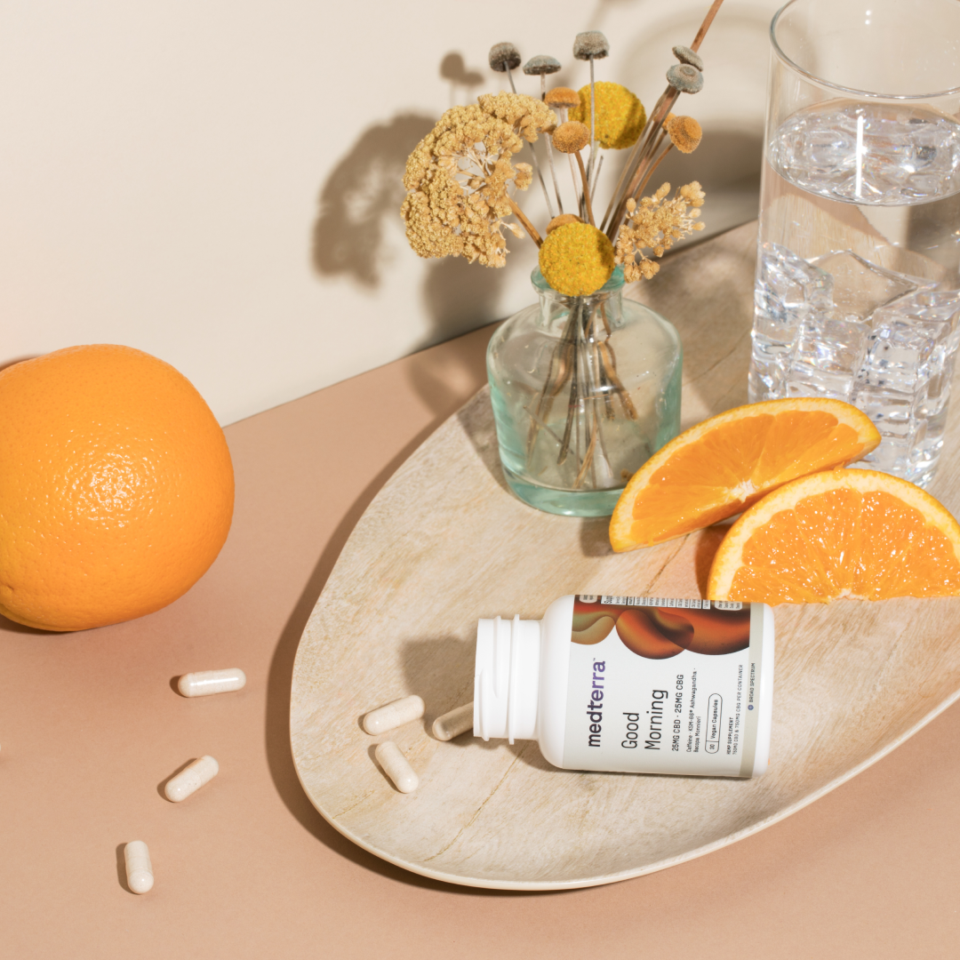 A tray displays a vitamin bottle labeled "Good Morning Capsules" from Medterra, surrounded by several capsules for cognitive support. The tray also holds a few orange slices and a half-full glass of water. A whole orange and a vase with dried flowers are in the background.