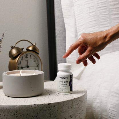 A hand reaches towards a bottle of "Good Night Capsules" by Medterra on a bedside table. Also on the table are a lit candle in a gray container and a brass-colored alarm clock showing 10:06. The background features white quilted fabric, setting the scene for this natural sleep solution featuring melatonin and magnesium.