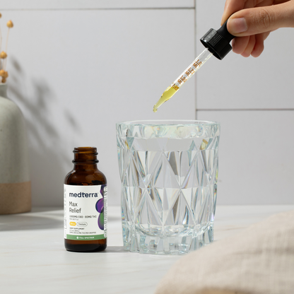 A hand is holding a dropper filled with yellow liquid over a glass tumbler filled with water. A brown dropper bottle labeled "Medterra Max Relief Full Spectrum CBD Oil"—an advanced tincture containing True Full Spectrum CBD from organically grown hemp—is placed beside the glass on a light-colored surface. The background includes a decorative vase.