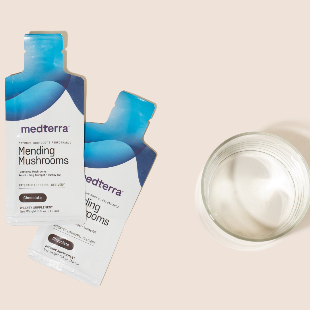 Two packets of Medterra Mending Mushrooms dietary supplement in chocolate flavor are displayed next to an empty, transparent glass bowl on a light beige surface. The blue and white packets feature text about the mushroom blend, highlighting its antioxidant benefits and support for athletic recovery.