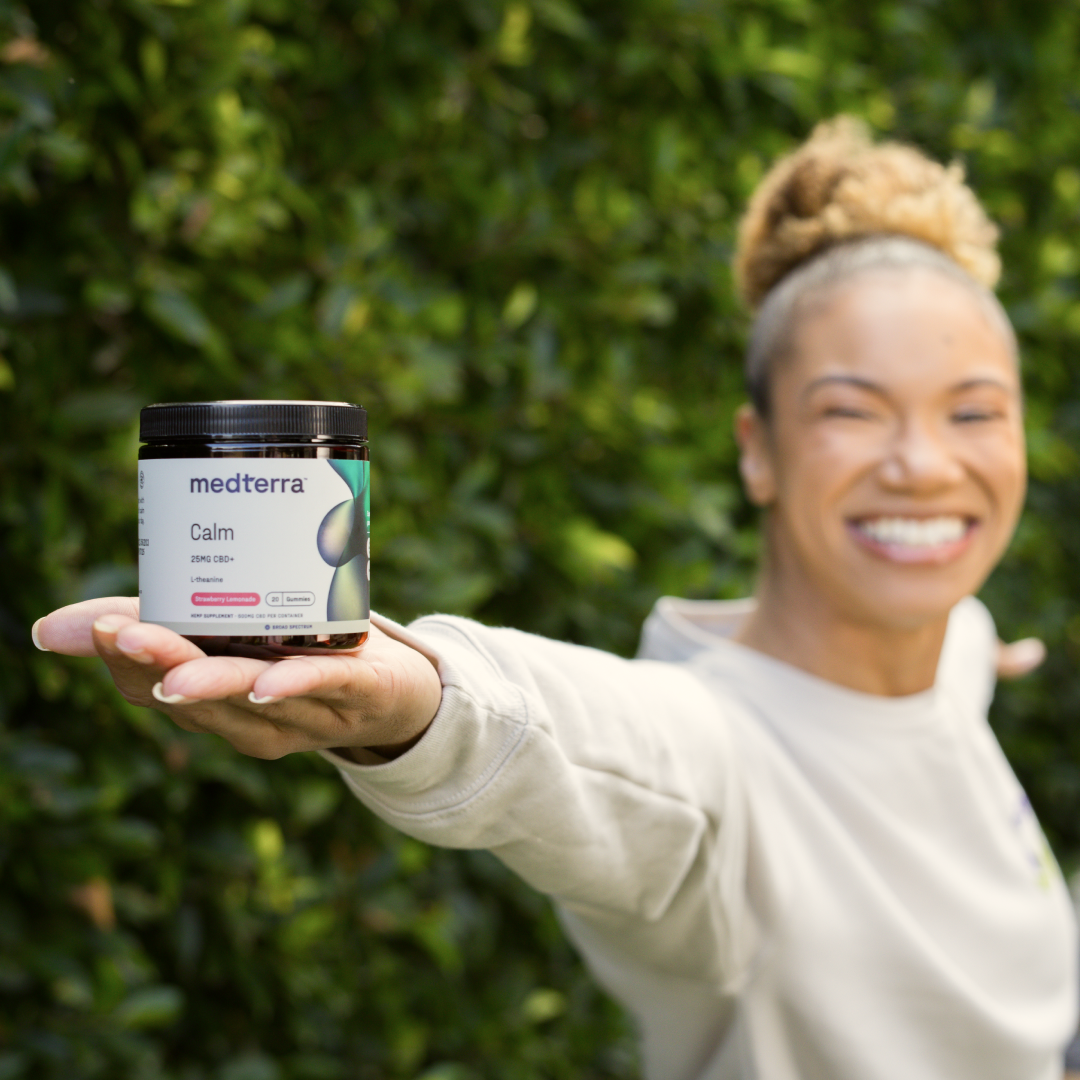 A cheerful individual with curly hair extends a jar of Calm CBD Gummies by Medterra towards the camera, showcasing the label that highlights Broad-spectrum CBD for stress relief. They are posed in front of lush green foliage, dressed in a light-colored long-sleeve shirt.