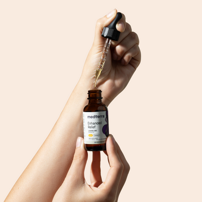 A person uses a dropper to extract liquid from a brown glass bottle labeled "Medterra Enhanced Relief Broad Spectrum CBD Oil." The bottle, likely containing Broad Spectrum hemp plant extract, is being held in one hand against a neutral, light-colored background.