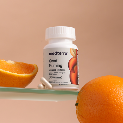 A bottle of Medterra Good Morning Capsules, designed for cognitive support and containing both CBD and CBG, rests on a glass surface with two capsules beside it. An orange slice and a whole orange are placed nearby. The background features a soft peach color.