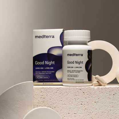 A white bottle labeled "Good Night Capsules" containing 50mg CBD and 1.5mg CBN by Medterra is placed next to its box on a textured, beige surface with abstract white decorations. Two beige capsules, part of this natural sleep solution from Medterra, are positioned beside the bottle.