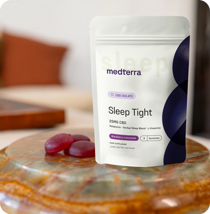 A bag of Medterra Sleep Kit sits on a table with two purple gummies beside it. The scene features a softly blurred background that includes part of a couch and a wooden surface.