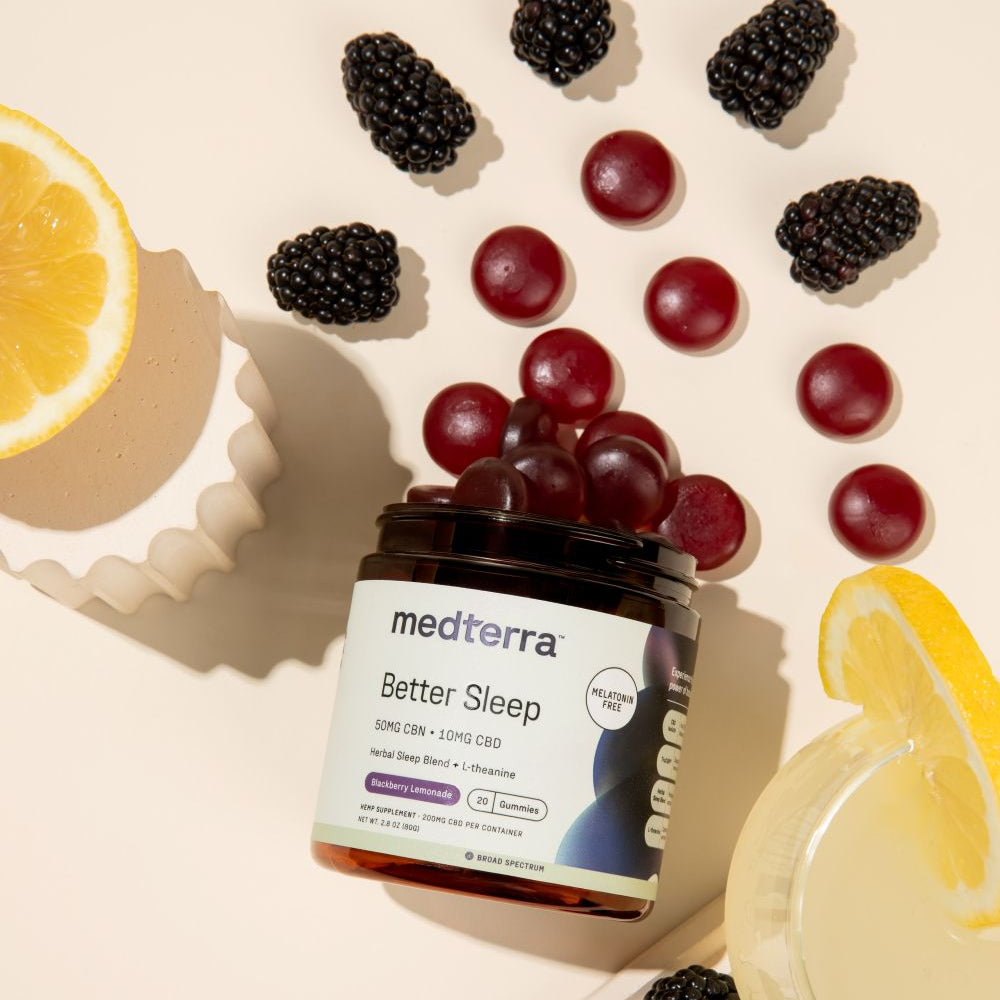 A jar of Medterra Better Sleep TruCBN® Gummies sits surrounded by blackberries, gummy pieces, lemon slices, and a glass of lemon water on a light background—showcasing these delicious TruCBN® CBD sleep gummies in a refreshing setting.