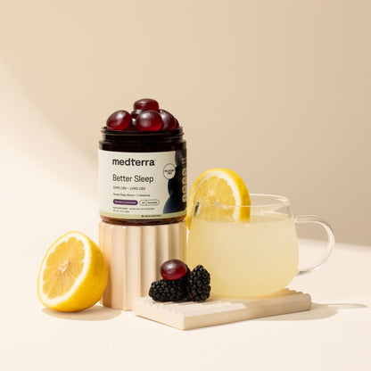A bottle of Medterra Better Sleep TruCBN® Gummies, a melatonin-free sleep aid, with red gummies, a glass of lemon drink, a half lemon, blackberries, and grapes arranged on a beige surface with soft shadows.