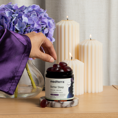 A hand in a purple sleeve picks up a Medterra Better Sleep TruCBN® Gummy from its jar on a wooden table, next to purple hydrangeas and three lit, cream-colored ribbed candles.