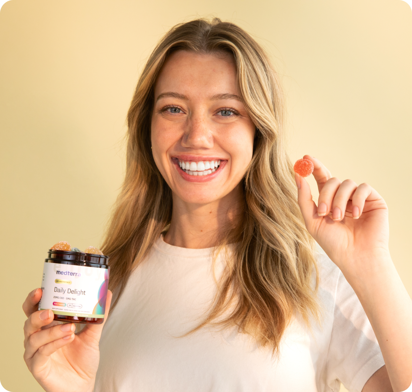 A smiling individual with long hair in a white shirt holds a gummy and a jar labeled "Delight & Good Night Bundle" from Medterra against a beige background.